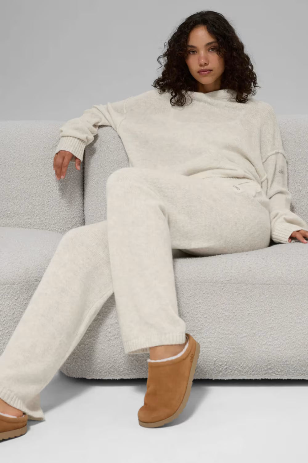 Mulher sentada em sofá usando chinelo bege de pelúcia com roupa confortável off-white, moda casual.