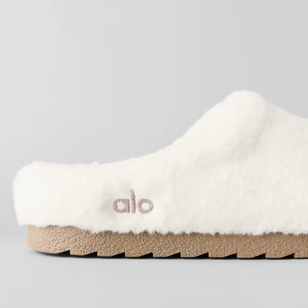 Slipper branco de pelúcia com sola bege, detalhe lateral alo, design confortável e moderno.