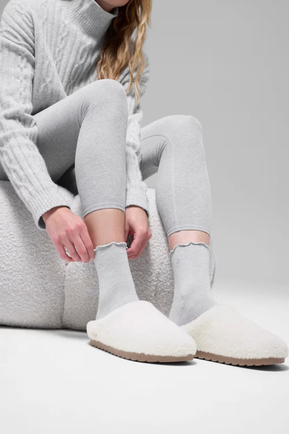 Modelo usando pantufa branca de pelúcia, meia cinza e roupa confortável, estilo casual.