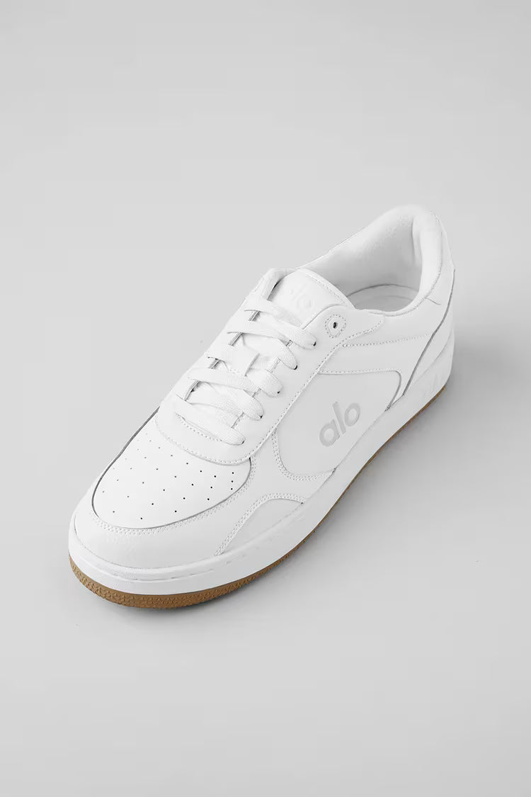 Tênis branco casual com sola marrom, detalhes perfurados e logo na lateral, fundo cinza.