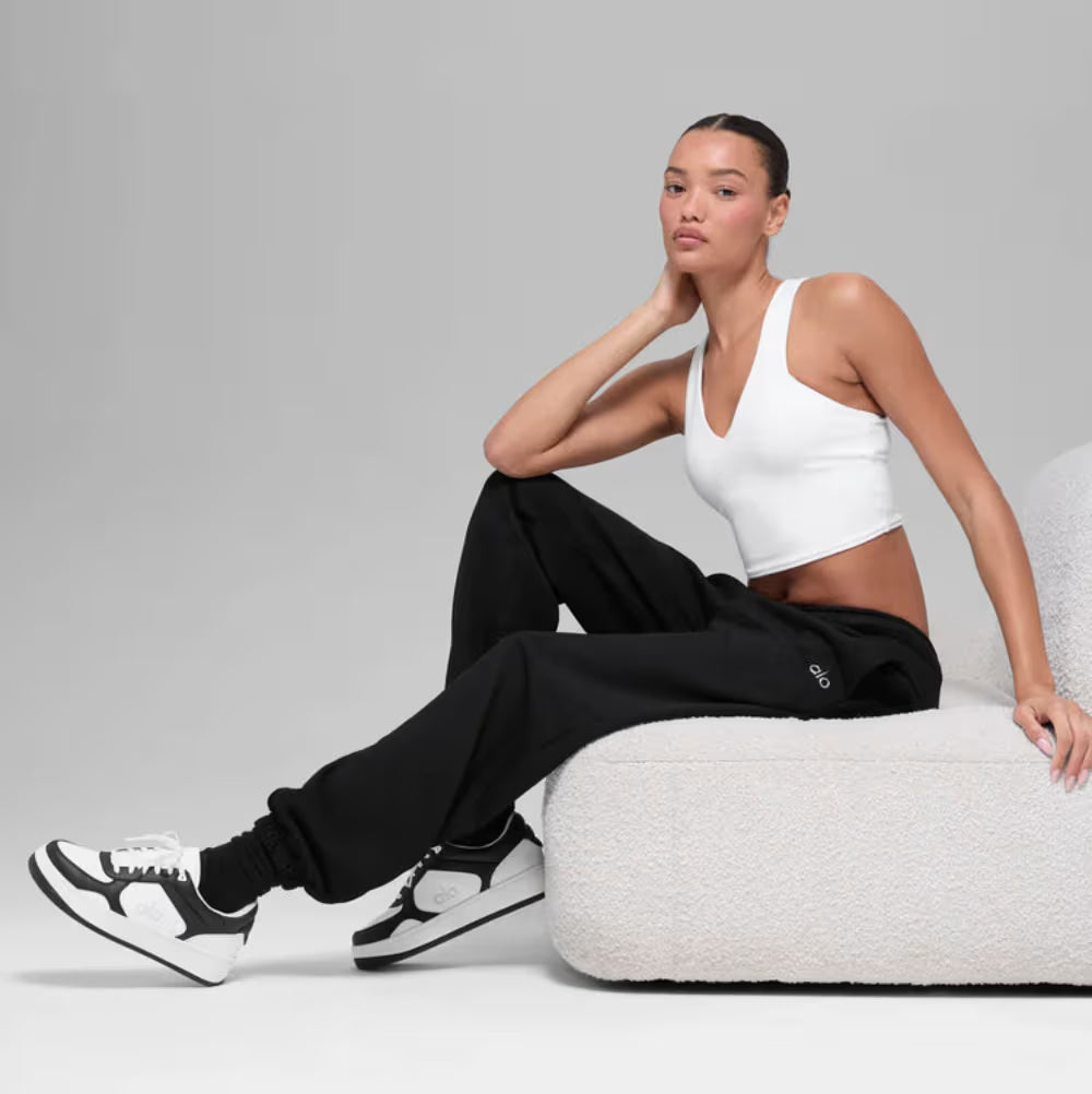 Mulher sentada em sofá moderno usando tênis preto e branco, calça jogger e top branco, estilo casual esportivo.