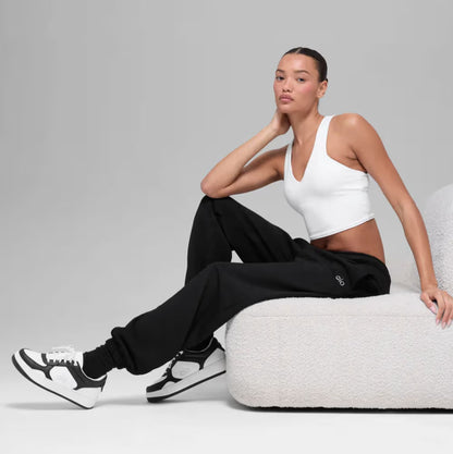 Mulher sentada em sofá moderno usando tênis preto e branco, calça jogger e top branco, estilo casual esportivo.