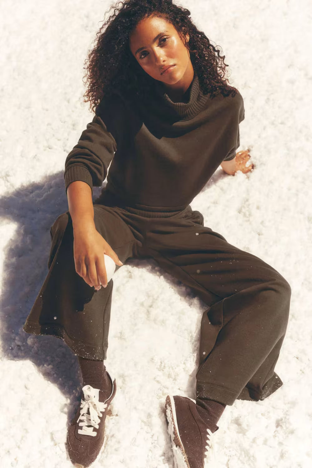 Mulher sentada na neve usando tênis marrom Alo Yoga Sunset e roupa casual escura.